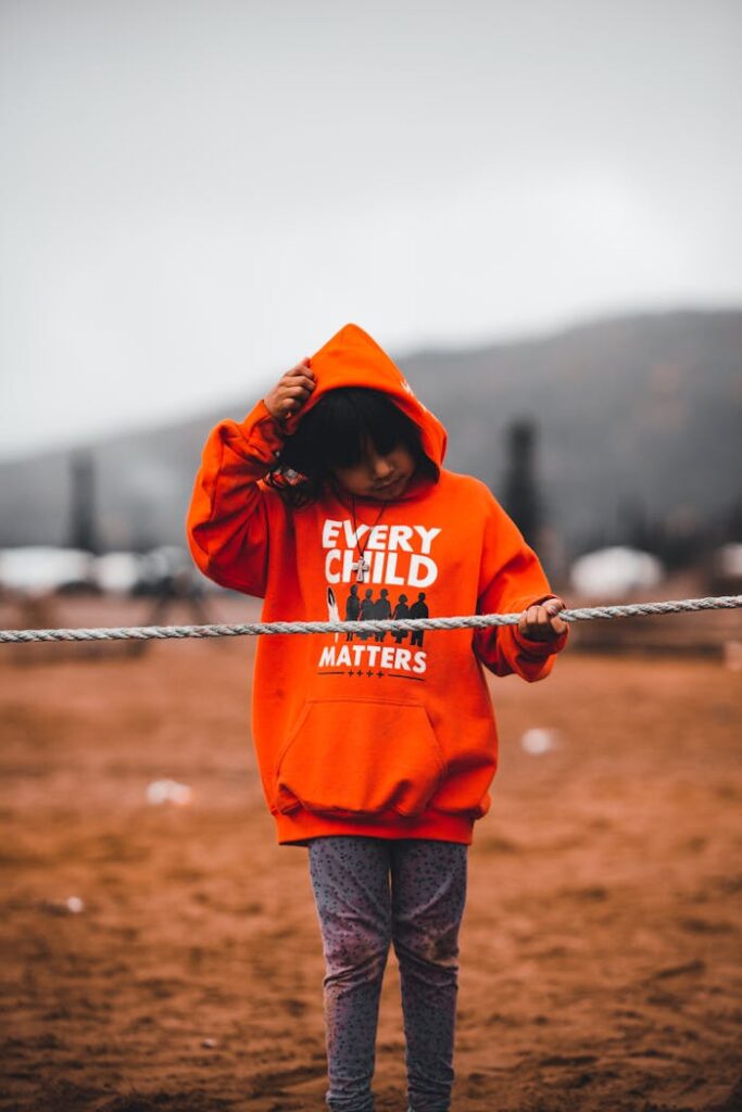 A child wearing an orange 'Every Child Matters' hoodie standing in a sandy outdoor area holding a rope.